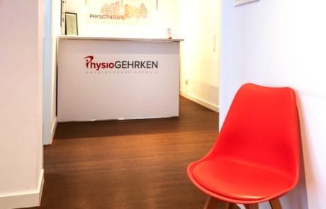 Roter Stuhl im Flur einer Physiotherapiepraxis mit einem Empfangstresen und einem Logo im Hintergrund.