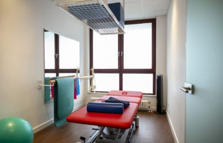 Ein Physiotherapieraum mit gepolstertem Behandlungstisch, an der Wand hängenden Gymnastikmatten, einem Gymnastikball und großen Fenstern.