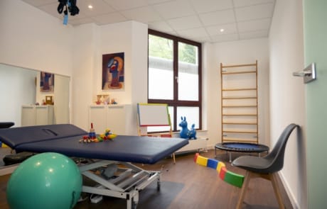 Ein Therapieraum mit einem verstellbaren Tisch, einem Gymnastikball, Spielzeug, einem Minitrampolin, einer Leiterwand und einem Stuhl. Der Raum hat ein großes Fenster und Holzboden.