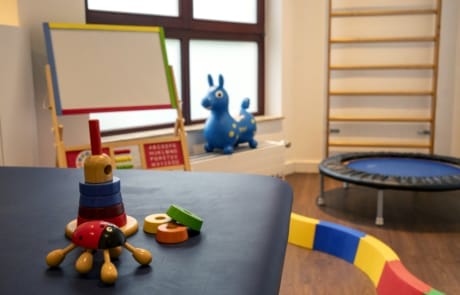 Ein Kinderspielzimmer mit stapelbarem Spielzeug auf einem Tisch, einer Staffelei, einem Trampolin, einem Schaukelpferd und Klettergerüsten.