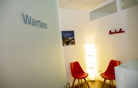 Ein kleiner, moderner Wartebereich mit zwei roten Stühlen, einer großen Lampe und einem Wandbild mit den Worten „Warten“ und „Lächeln“.