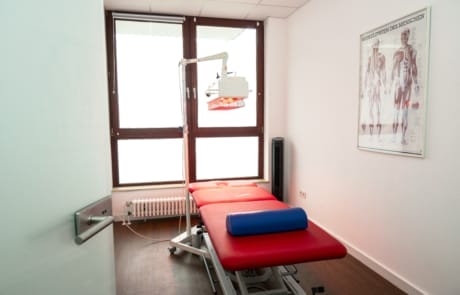 Ein medizinischer Untersuchungsraum mit einem roten Untersuchungstisch, einer medizinischen Deckenlampe, einem anatomischen Poster und großen Fenstern.