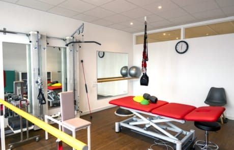 Ein Physiotherapieraum mit einem roten Behandlungstisch, Fitnessgeräten, Gymnastikbällen, einem Stuhl und einer Wanduhr.