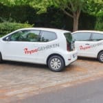 Zwei weiße Autos parken auf einer Straße, beide sind mit der Aufschrift „Physio Gehrken“ in roter und schwarzer Schrift versehen. Im Hintergrund sind Bäume und Laub zu sehen.